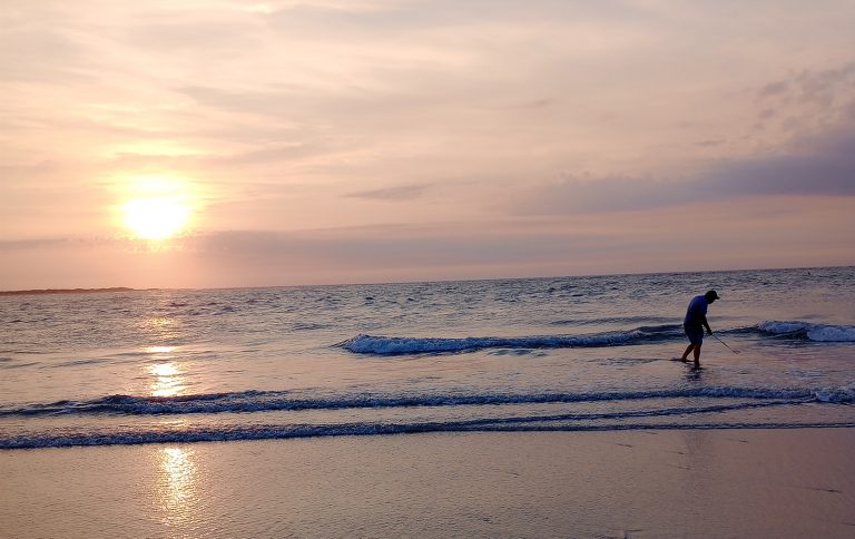 A man shelling in shallow beach water during sunrise